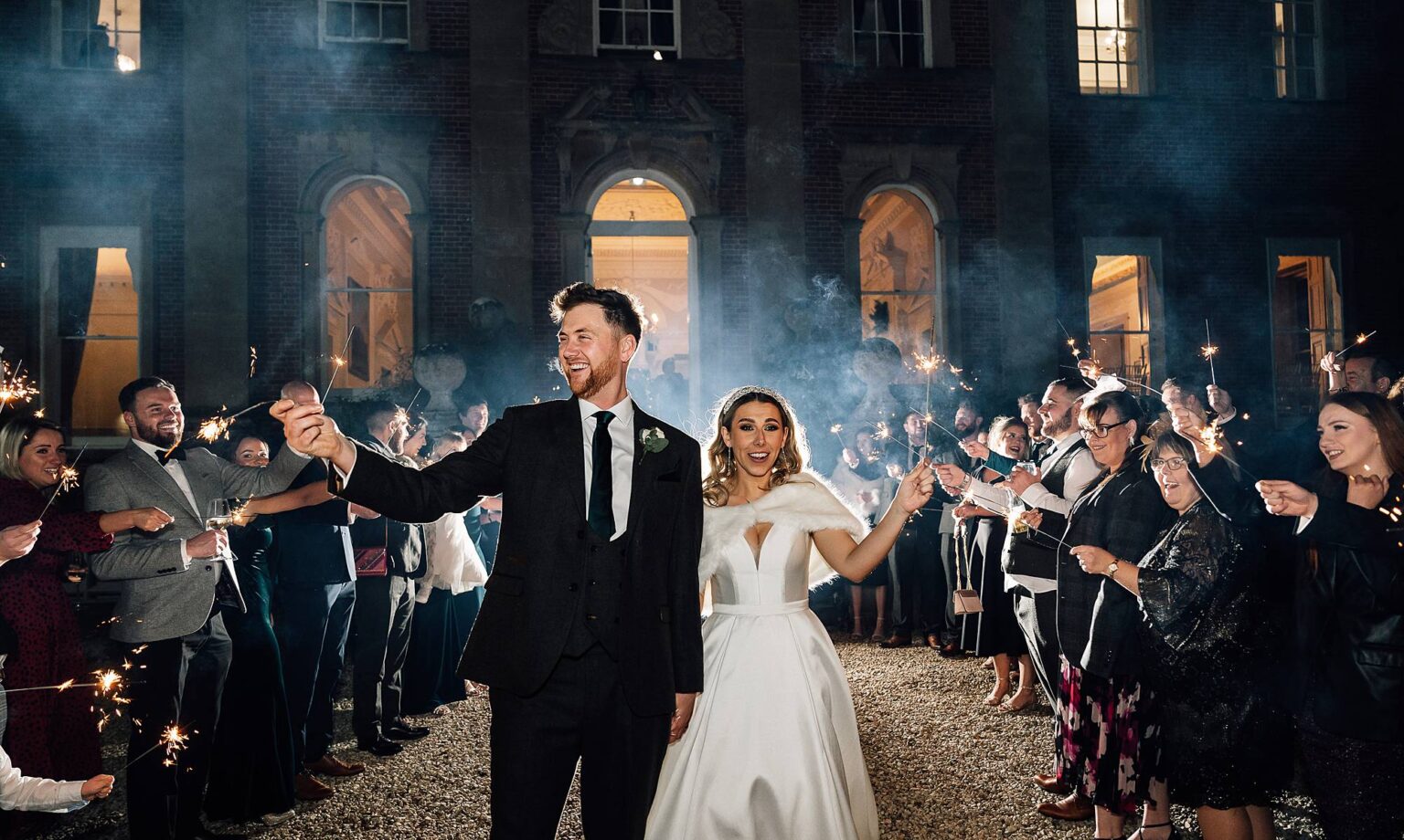 Bride and groom walk down a sparkler tunnel with guests either side of them and Crowcombe Court behind